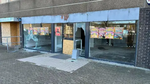 A vandalised shop front, with smashed windows and doors.