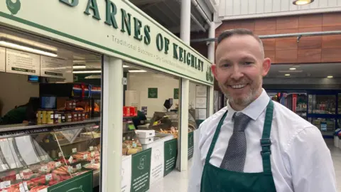 A man in white shirt and tie and green apron standing outside a butcher's shop with a yellow and green sign above it
