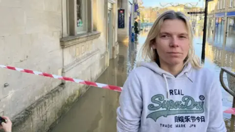 Marie Indge/BBC A blonde-haired woman wearing a hoodie stands in front of caution tape in front of a flooded street.