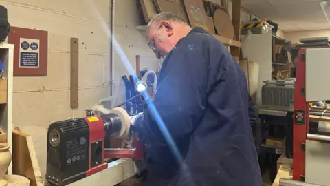 Isabella Verona/BBC A man working at a woodturning lathe inside a workshop. The lathe is spinning a piece of wood while the person leans forward, shaping it. A bright task light shines directly onto the work area, creating a visible beam of light. The surrounding area includes shelves with tools, framed items, and workshop equipment. 