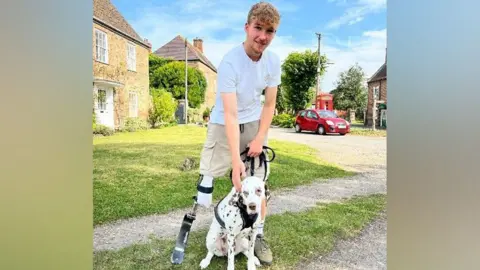 Ollie standing with his prosthetic blade alongside a Dalmatian dog on a lead in a village setting on a sunny day