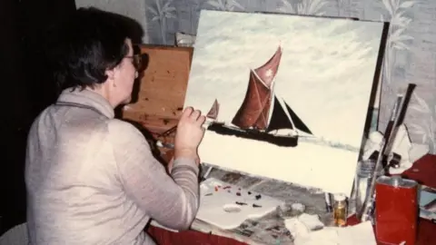 David Gilbey A woman painting a sail boat onto a canvas. She has her back turned to the camera and is wearing a beige jumper.