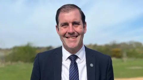A smiling Lee Dillon, the MP for Newbury, wearing a dark blue suit, white shirt and striped blue and gold tie. He's standing in open green fields with a low hedge behind him.