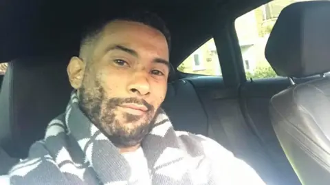 Metropolitan Police Leroy Mitchell is sitting in a car, smiling into the camera in a selfie-style photo. He has short black hair and a short black beard and is wearing a black and white scarf. 