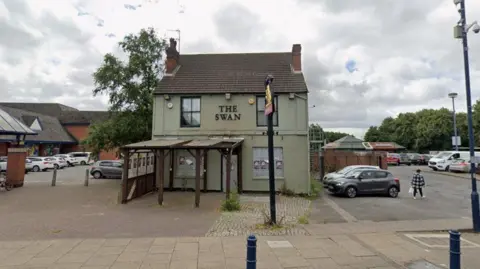 A pub building painted in grey, surrounded on three sides by a retail car park. "The Swan" is spelled out in black letters on the front of the building, and there is a gold, black and red Banks's Brewery sign outside.