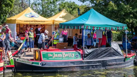 Lichfield and Hatherton Canals Restoration Trust A traditional narrowboat named Montgomery, carrying a sign for The Jam Butty preserves, is moored beside a towpath. Yellow and turquoise gazebos are set up behind it, one branded for the Lichfield and Hatherton Canals Restoration Trust. Visitors browse the stalls in bright sunshine.