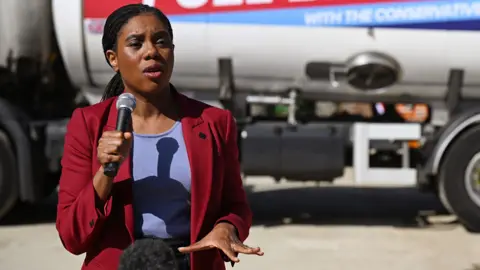 Badenoch speaks with a microphone in hand and gestures with the other hand while wearing a suit jacket and standing in front of a fuel tank truck with a Tory slogan on the side (the words 'with the Conservatives' are just legible in frame), in London last week.