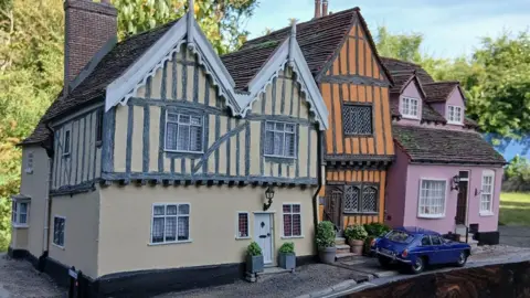 Simon Evans A model of a row of three cottages. There is one large cream cottage at the left end, in the middle is an orange smaller cottage and on the right end there is a pink cottage. Two of the homes look like medieval style buildings and there is a blue car model parked outside of the homes. 
