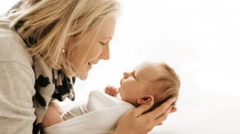 CLAIRE LONG Sophie, who has a blonde bob and wears gold drop earrings with a grey jumper and a grey, pink and black scarf, cradles her new-born daughter Martha, who is dressed in swaddling clothes