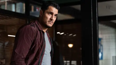A South Asian man wearing a burgundy jacket and grey t-shirt stands in an office with shuttered blinds. An office setting is behind him in the photo.
