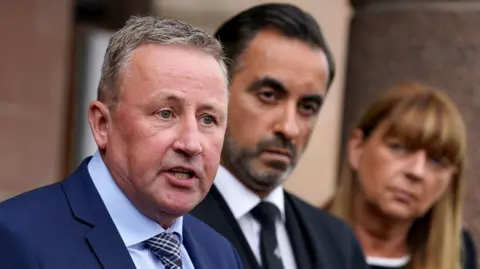 Stuart Allan has short grey has and wears a navy suit. He is speaking and stands next to Aamer Anwar, a man with short dark hair and trimmed grey facial hair. Next to him is Linda Allan, who has shoulder length brown hair who is watching her husband speak.