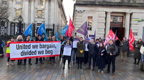 People gathered outside High Court in Chichester Street, Belfast, County Antrim, on Friday. They hold blue, red and purple flags and a sign that says 'United we bargain, divided we beg'.