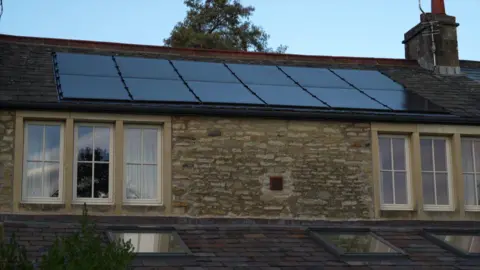 A row of nine black solar panels on the roof of a country cottage style house.