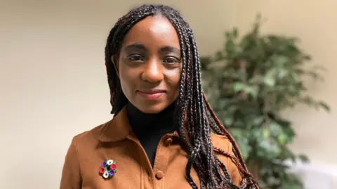 Councillor Hana John is smiling for the picture. She has long, braided hair and is wearing a tan colour jacket, with a flower broach. 