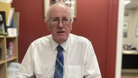 Jim Shannon, a man with grey hair and glasses, wearing a white shirt and blue tie.