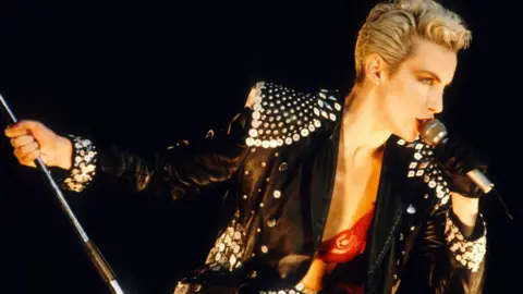 Getty Images Annie Lennox singing into a mic during the 1980s. She has short, cropped, blonde hair and is wearing a black leather jacket with sequins attached