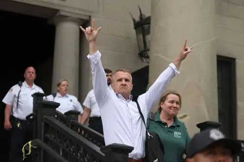 Boston Herald via Getty Images Aidan "Turtleboy" Kearney, wearing a white shirt gestures outside the courthouse. His hand gesture means "I love you" in American Sign Language
