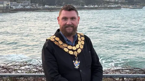 Drew has brown hair and beard and wears a dark jacket and blue shirt and has his mayoral chains around his neck. He stands with the sea behind him. 
