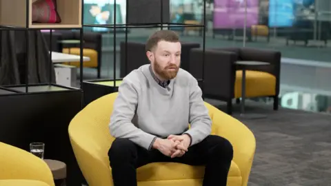 Nye Jones from Generation Rent is sitting in a large open plan office space on a bright yellow chair. He has his hands crossed and is talking to somebody out of view