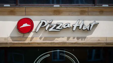 A Pizza Hut restaurant logo that features white letters against a beige background and a red dot with a white hat in the middle. 