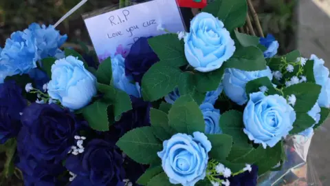 A blue flower arrangement is among the tributes with a card fastened to it, that says "RIP, love you cuz".