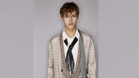 Dior Guernsey sixth form student Jack Paulsen modelling for fashion brand Dior in Paris. He is looking at the camera and is wearing a jacket, , a white shirt, an embroidered tie and a very thin scarf over his shoulder. 