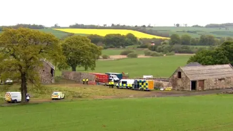 Emergency vehicles and police tape at some old farm buildings surrounded by fields