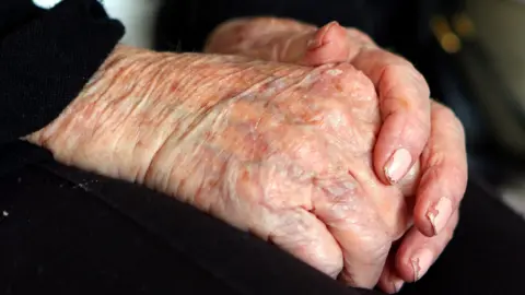 An elderly woman's hands, one placed on the other, in her lap. She is wearing black trousers and has chipped pink nail varnish on her fingernails. 