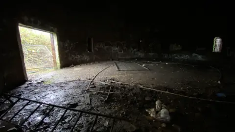 Zac Sherratt/BBC A dark room with mud and various pieces of metal on the floor. There is a large square hole in the left side of the building, which is letting in light