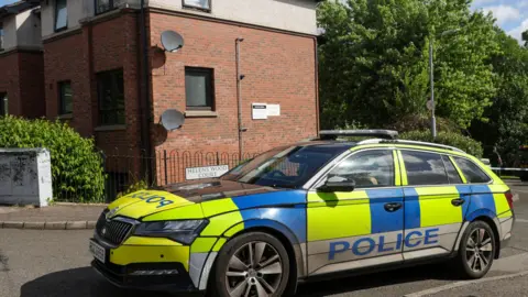 A marked yellow and blue police car parked outside an apartment block. There are trees with green leaves in the background and it appears to be a bright day.