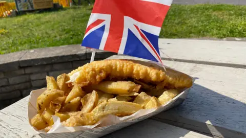 A portion of fish and chips in a cardboard tray with a union flag tucked in beside it