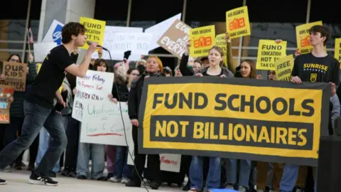 A young man speaks into a microphone while facing a group of protesters holding signs. A large yellow and black sign reads "Fund Schools Not Billionaires"