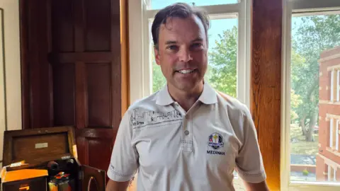 Ken Skates standing in front of a large window inside a wood-panelled room. The person is wearing a white polo shirt featuring the Ryder Cup 2012 "Medinah" logo. Beside him is a wooden display cabinet containing decorative items, while trees and a red-brick building are visible through the window behind.