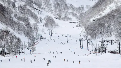 Skiers dot a snowcovered mountain slope with cable cars and trees at the sides.