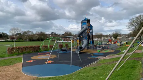 Torridge District Council Play park with a tall structure with a slide. There is other equipment including swings and climbing frame in the centre of the park on Tarmac next to a big field.