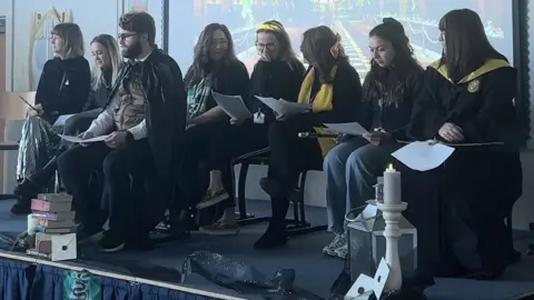 Elliot Deady/BBC Eight teachers sitting on a stage at the front of a school hall. They are all wearing Harry Potter-themed fancy dress.