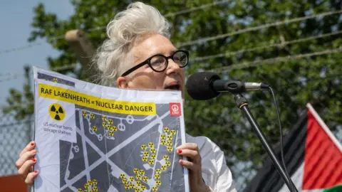 A woman with white hair and dark glasses holding up a placard showing potential nuclear weapons storage sites at RAF Lakenheath