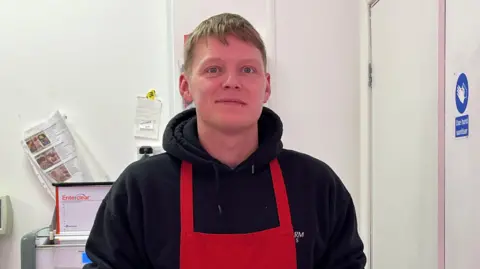 Butcher Wayne Darbyshire with blond hair and a lip ring wearing a black hoodie and red apron. He stands behind the counter in a butchers shop. He is smiling.