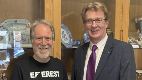 Two men standing side-by-side smile at the camera. The man on the left is wearing a black top and the man on the right is wearing a suit and tie. They are standing in front a display cabinet which contains a number of plates. 