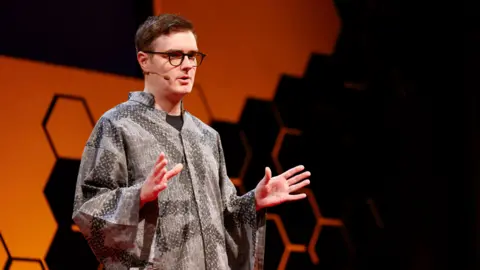Ted AI Welsh computer scientist Llion jones stands on stage at a TED-AI conference in San Francisco, wearing a grey patterned, loose-fitting shirt. The background features a bold orange shape with a honeycomb-style black geometric design. Llion wears glasses and a microphone and is mid-presentation, with both hands raised in a gesturing position.