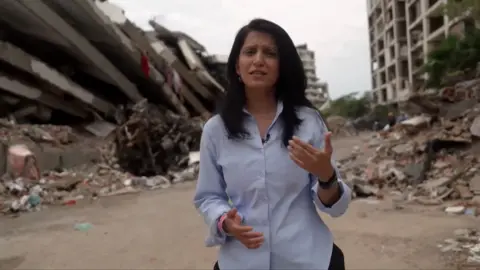 Yogita wears a blue shirt and speaks to the camera as she stands in front large piles of rubble, including what looks like a collapsed apartment block.
