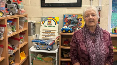 LDRS Dot Capes wearing a purple top and scarf, standing alongside shelves of toy cars.