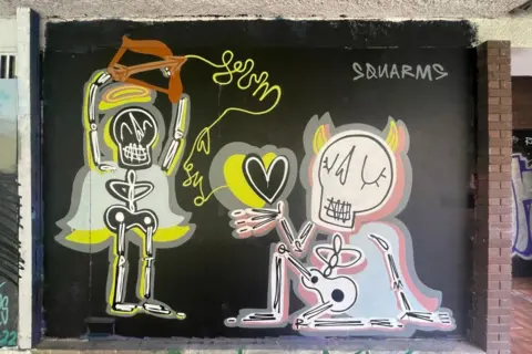 James Thompson/Wall Games A street art mural featuring two cartoon skeletons, one is shooting an arrow from a bow into a love heart being held by the other.