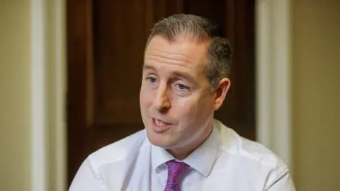 A man with grey hair is wearing a white shirt and pink neck tie. He is speaking at the camera. Behind him is a wooden door.