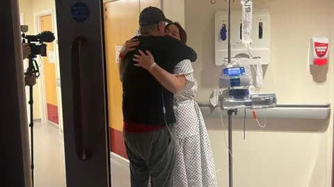 John and Lorna, who is wearing a hospital gown, hug in a corridor ahead of the surgery. 