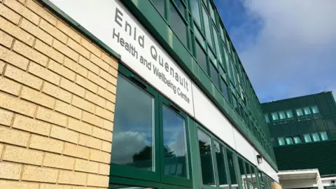 A view from the ground up of a tall, green building. It has lots of windows. A white sign above the first row of glass says 'Enid Quenault Health and Wellbeing Centre'. At either ending of the building, the ground floor is built with a column of light brown bricks.