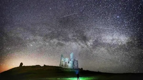 Grey castle ruins are illuminated by thousands of stars in the sky. The milky way is visible, as are shooting stars. In the foreground, there is a silhouette of a man looking up at the sky with a torch.