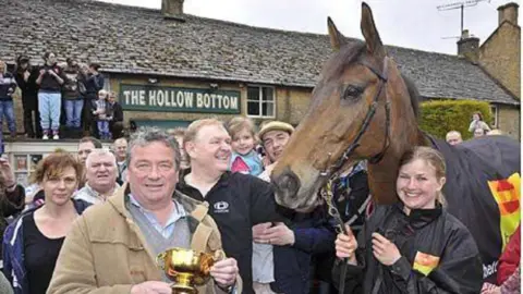 A large crowd stands outside of The Hollow Bottom - a traditional Cotswold pub - celebrating a Gold Cup win. People can be seen standing on a flat top roof above the door. One man at the front of the crowd holds a gold cup while a girl holds the winning horse's reins.