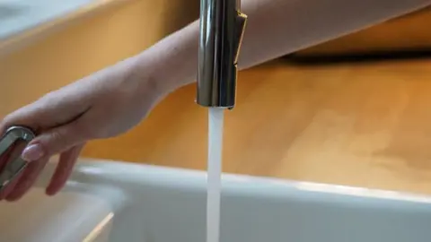 BBC A person with their hand on the tap as water runs out into the sink