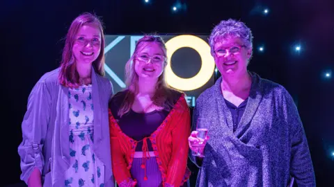 Kent County Council Three women illuminated with blue and purple lights in a dark room. They are looking at the camera and smiling. The woman on the right of the image is holding a champagne flute.